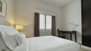 A hotel room with a bed, a desk, a chair, a lamp, and a window with curtains.