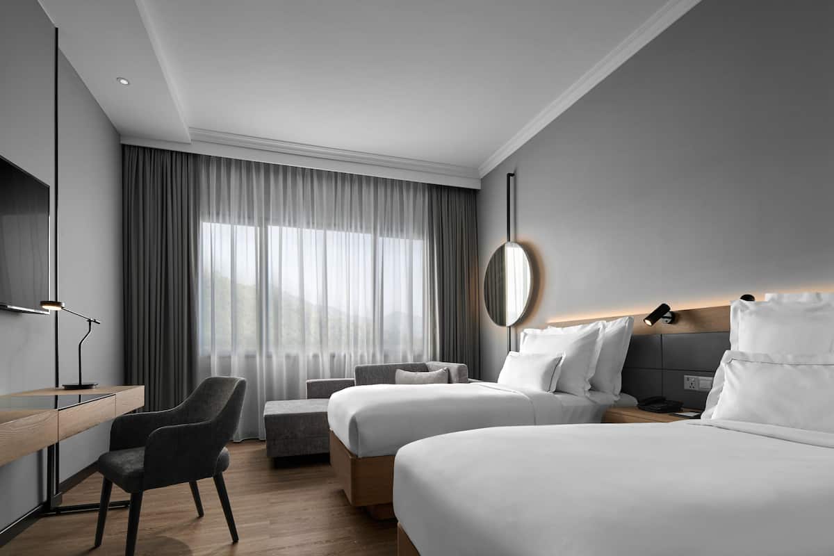 A modern hotel room with two beds, a desk, a chair, and a large window with curtains.