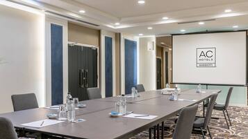 A conference room with a long table, chairs, water bottles, and a projector screen displaying the AC Hotels logo.