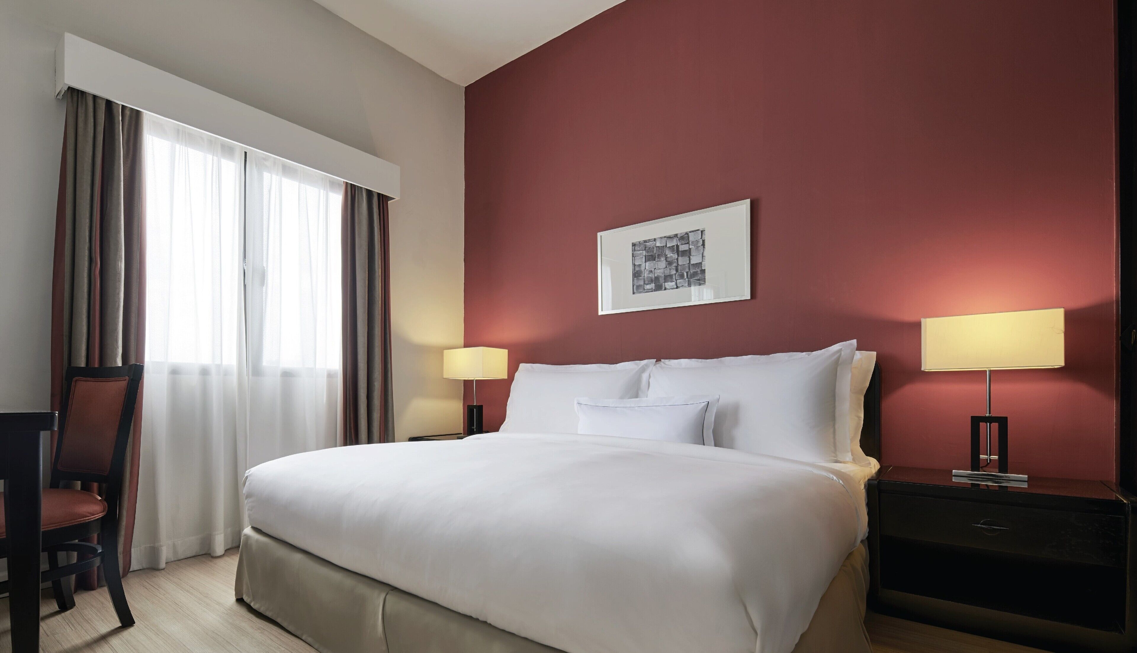 Una camera d'albergo con un letto grande, due comodini con lampade, una sedia, una finestra con tende e un quadro appeso al muro.