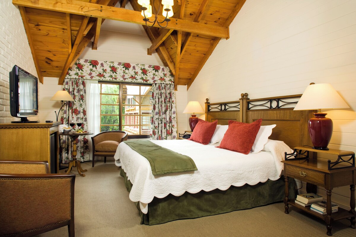A cozy bedroom with a wooden ceiling, a bed with white bedding and red pillows, a desk with a lamp, and a television.