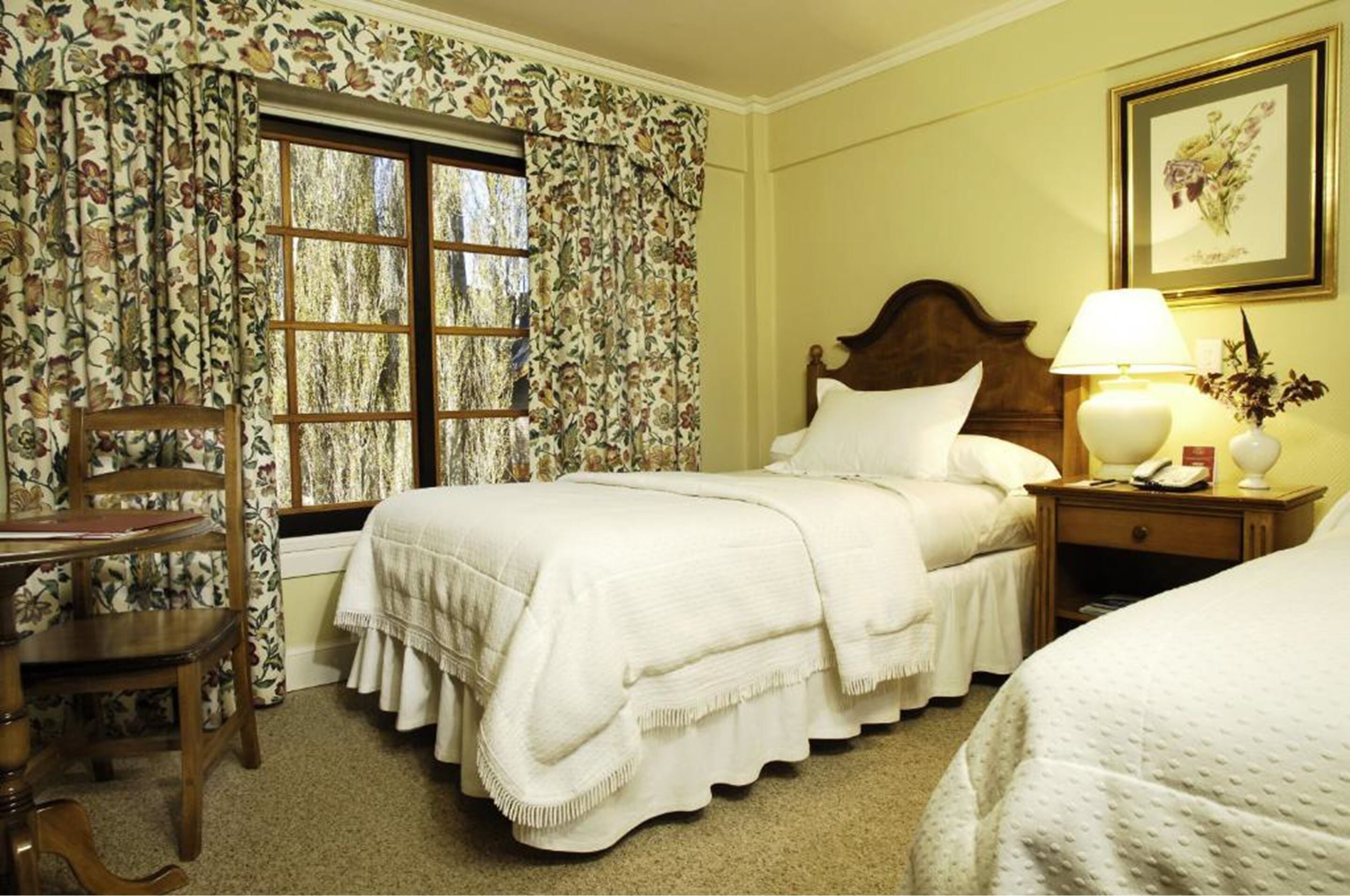 A neatly arranged bedroom with a bed, a chair, a nightstand with a lamp, and a window with floral curtains.