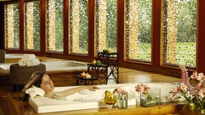 A spa room with a bathtub, a wooden floor, and large windows with a view of trees. There are candles and a vase with flowers on the windowsill. - Hotel Posada Los Alamos (El Calafate)