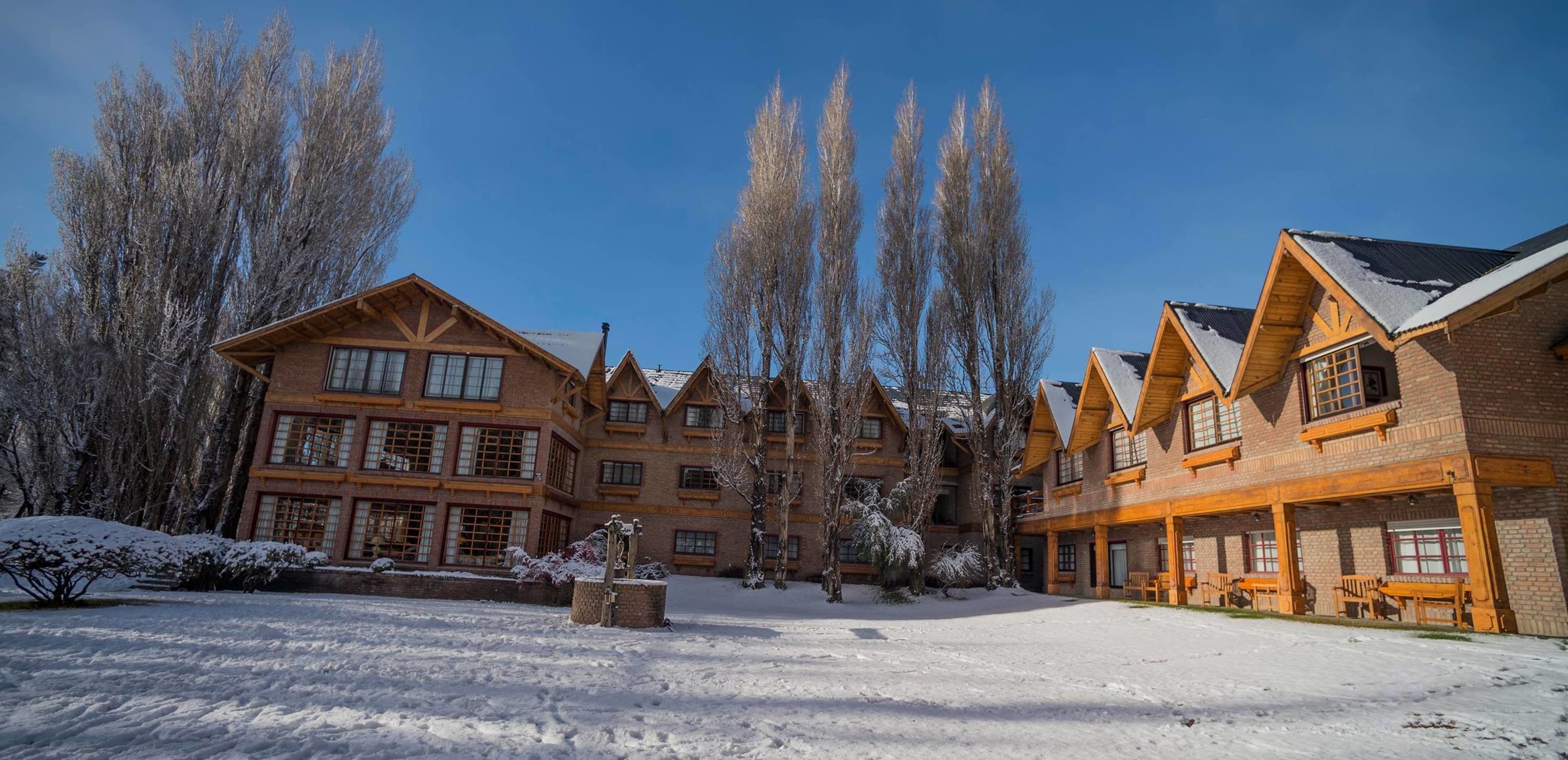 Una cabaña con varios edificios de madera, un patio cubierto de nieve y altos árboles.