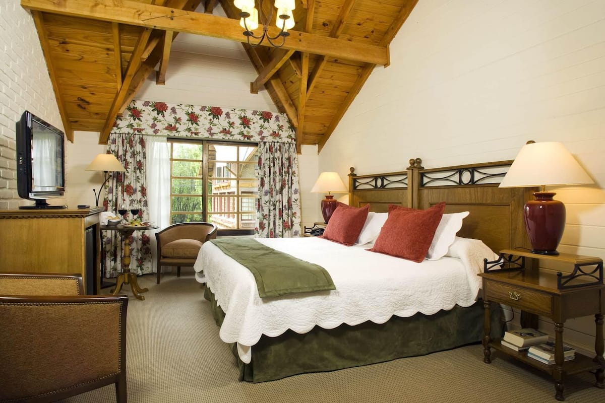 A cozy bedroom with a bed, two bedside lamps, a television, a chair, and a window with floral curtains.