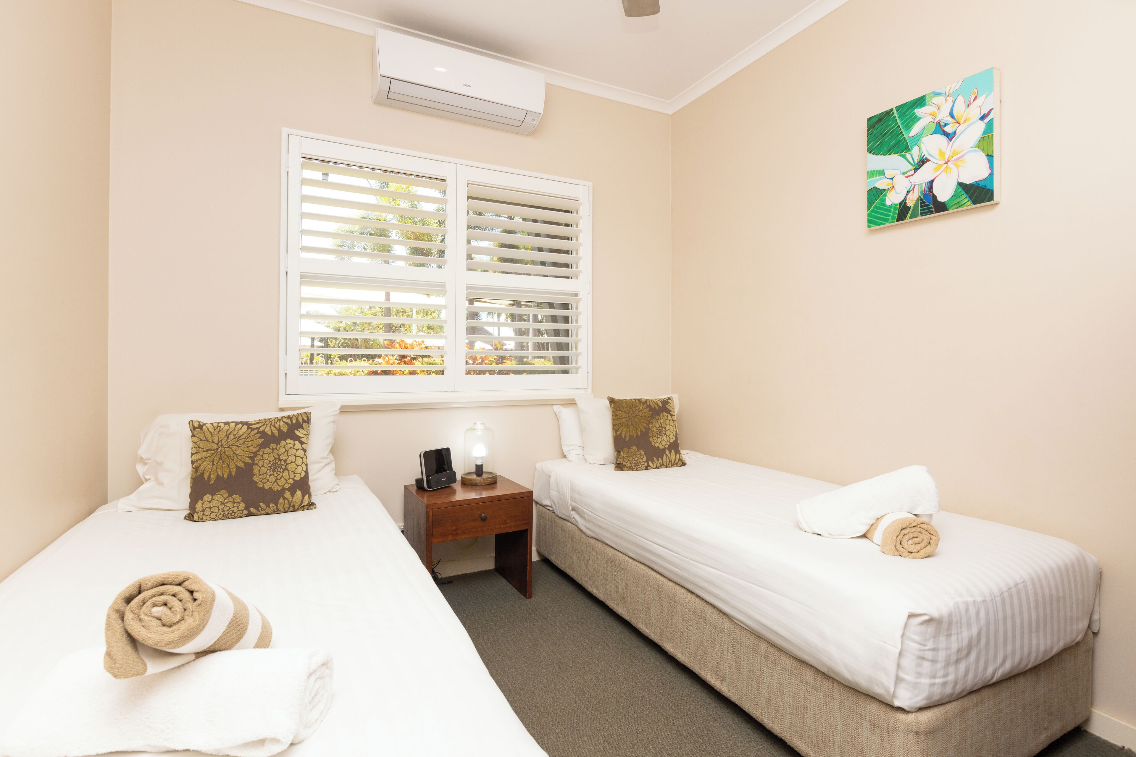 A hotel room with two single beds, a nightstand, a lamp, and a window with blinds.