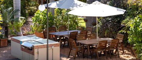An outdoor patio with a brick floor, wooden tables and chairs, and a built-in grill.