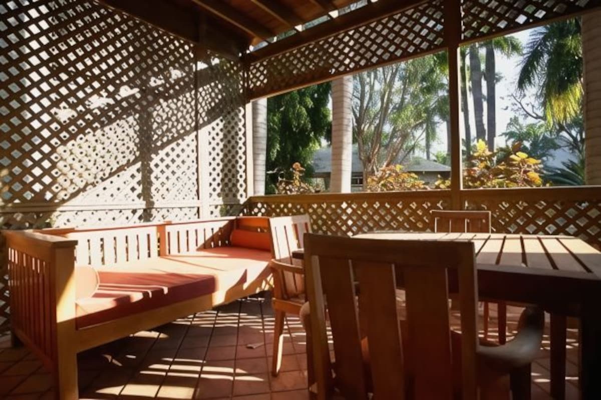 A covered outdoor seating area with wooden furniture and a view of palm trees.