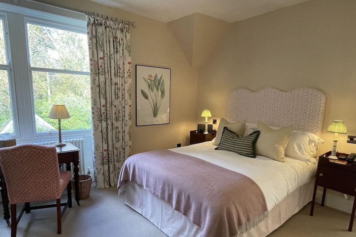 A bedroom with a large bed, two bedside tables with lamps, a chair, a window with curtains, and a framed botanical print on the wall.