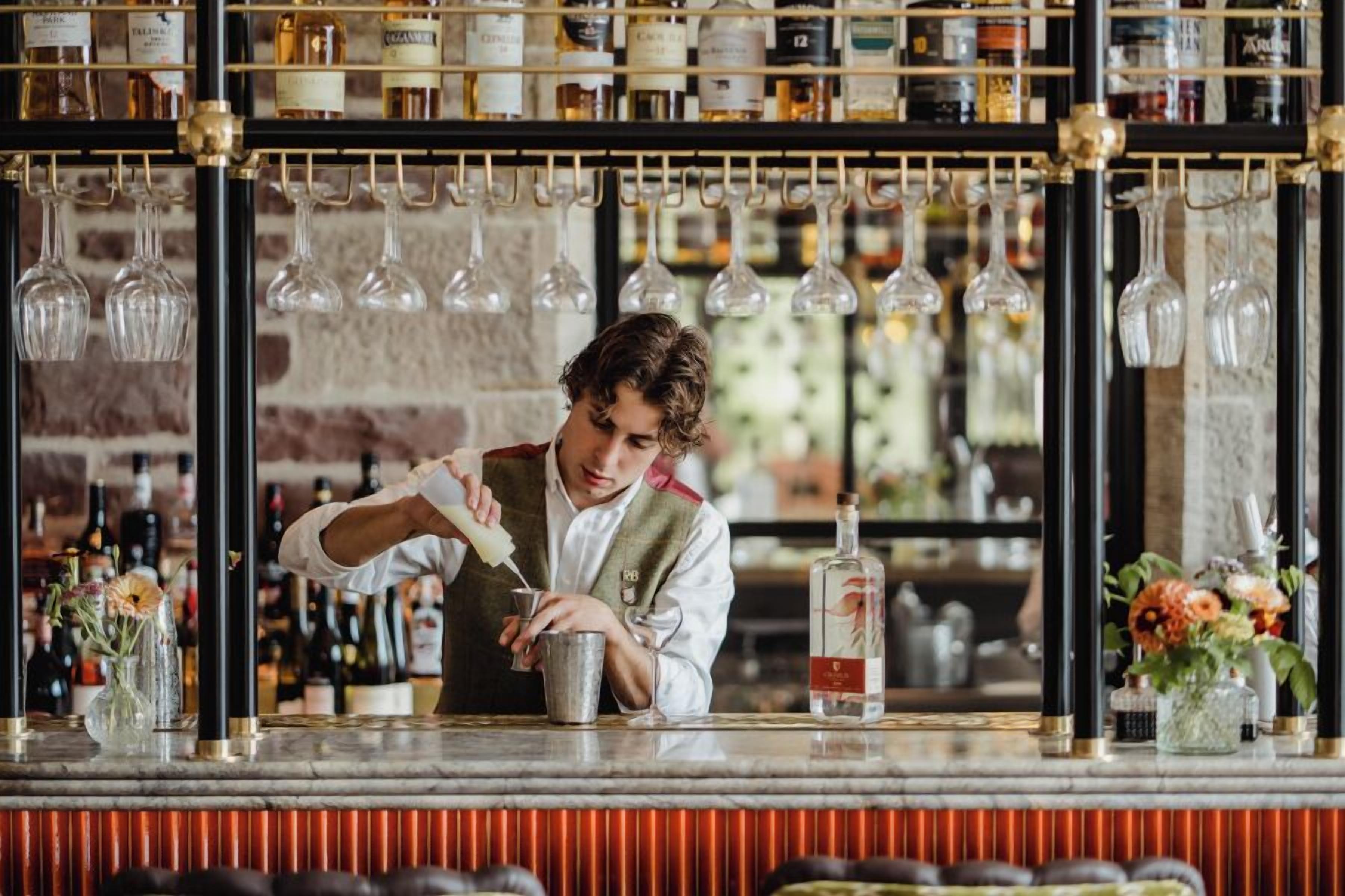 A bartender preparing a drink at a bar with a wine rack in the background.