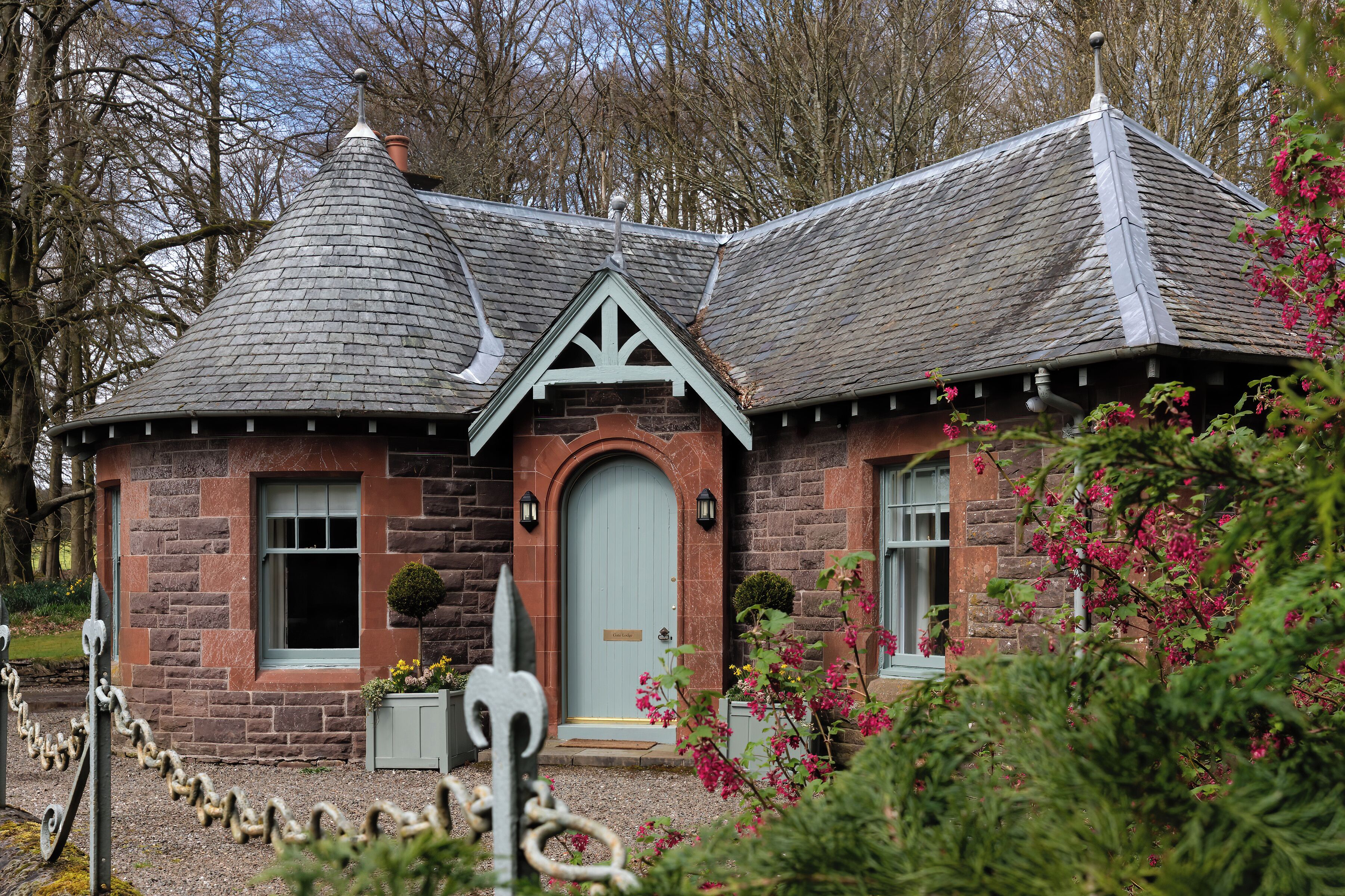 A stone house with a distinctive conical roof, a central door, and a small garden with pink flowers.