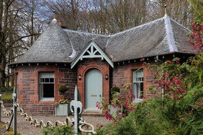 A stone house with a distinctive conical roof, a central door, and a small garden with pink flowers.