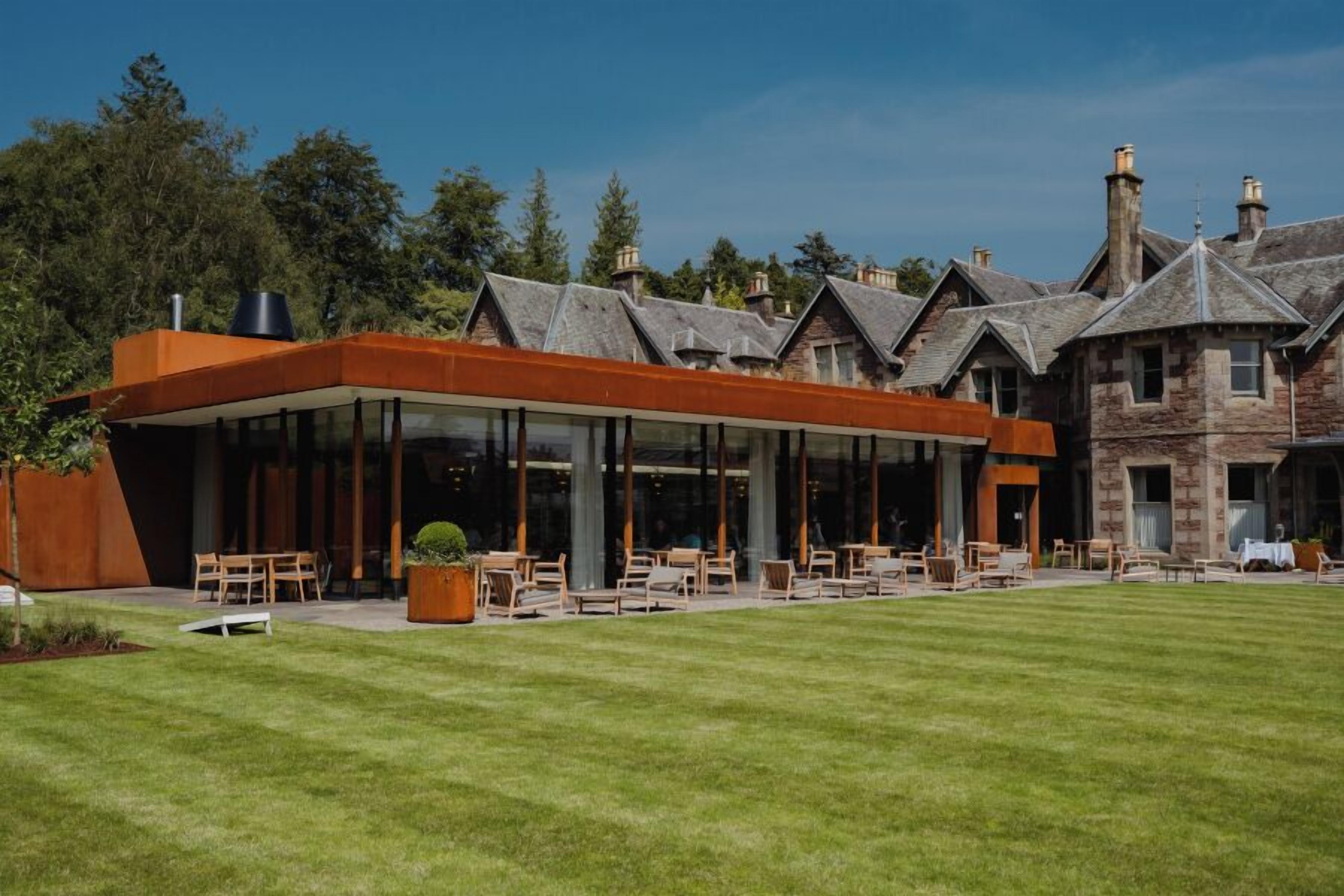 A modern building with a glass facade, outdoor seating area, and traditional stone buildings in the background.