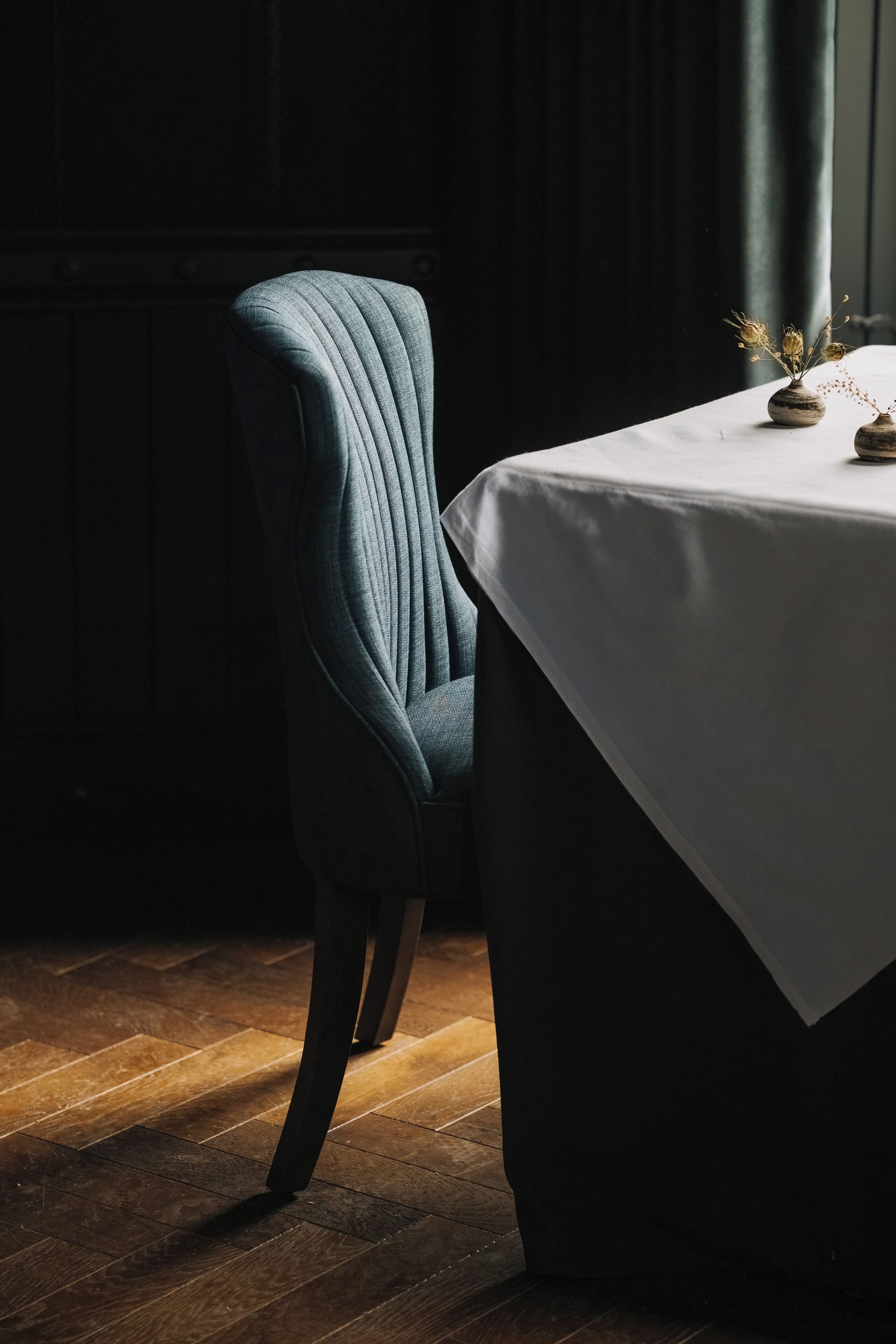 Un comedor con una mesa preparada para dos, una silla y un jarrón con flores secas.