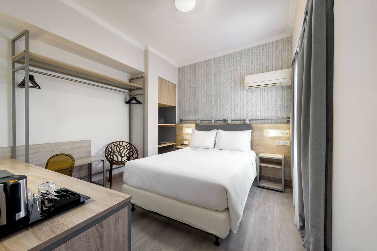 A hotel room with a bed, a desk with a coffee maker, a chair, a wardrobe, and a small table.