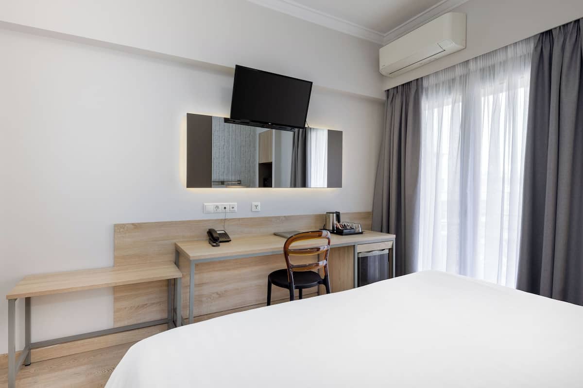 A hotel room with a bed, a desk with a chair, a flat-screen TV mounted above the desk, and a window with curtains.