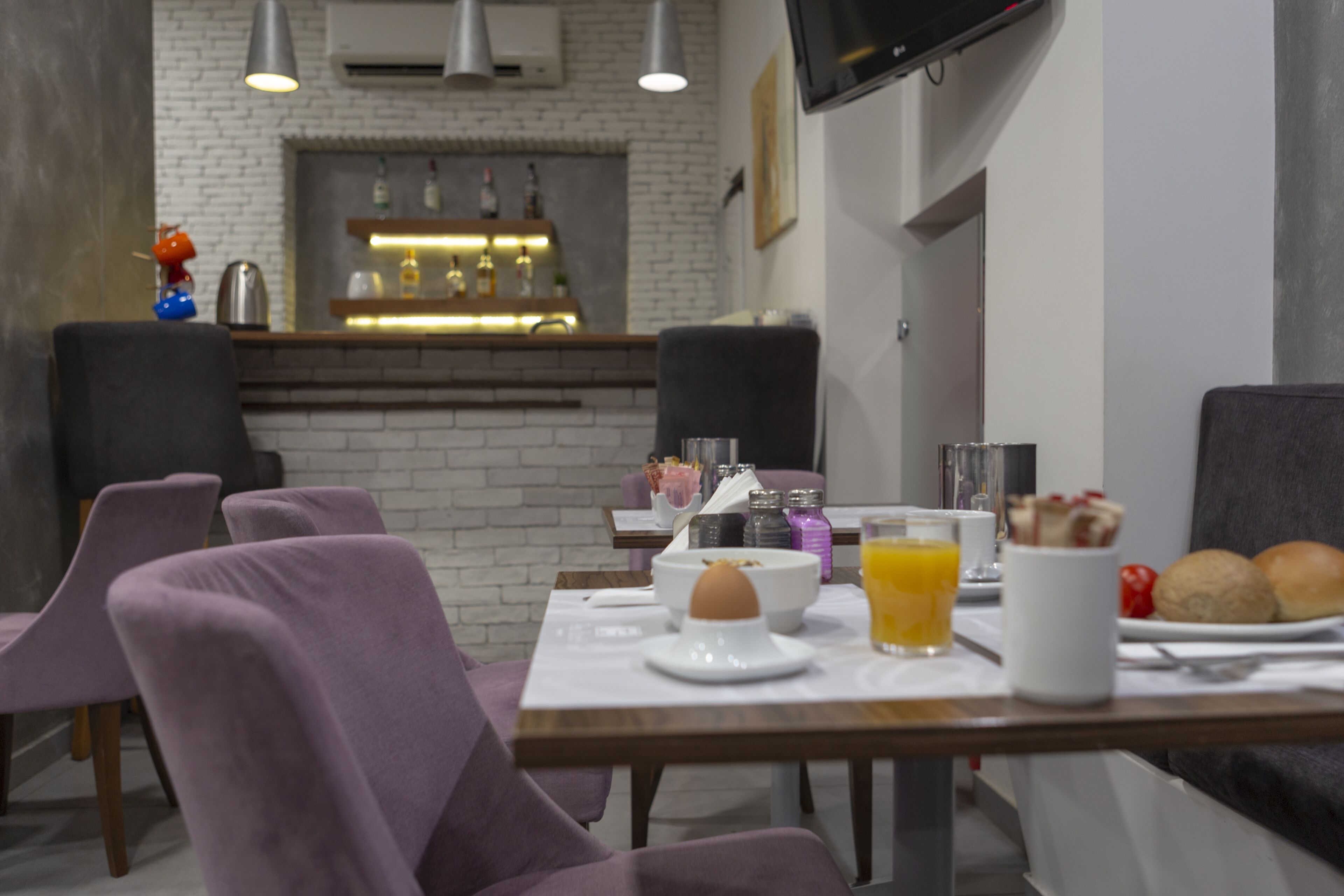 A modern dining area with purple chairs, a breakfast table with eggs, juice, and pastries, and a bar area in the background.