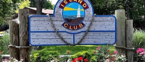 Une enseigne en bois portant l’inscription « ISLAND CLUB » et un emblème circulaire représentant un phare et des vagues.
