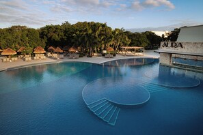 Une piscine dotée de chaises longues et de parasols en chaume, entourée de palmiers et d’un bâtiment portant l’inscription « AURA ».