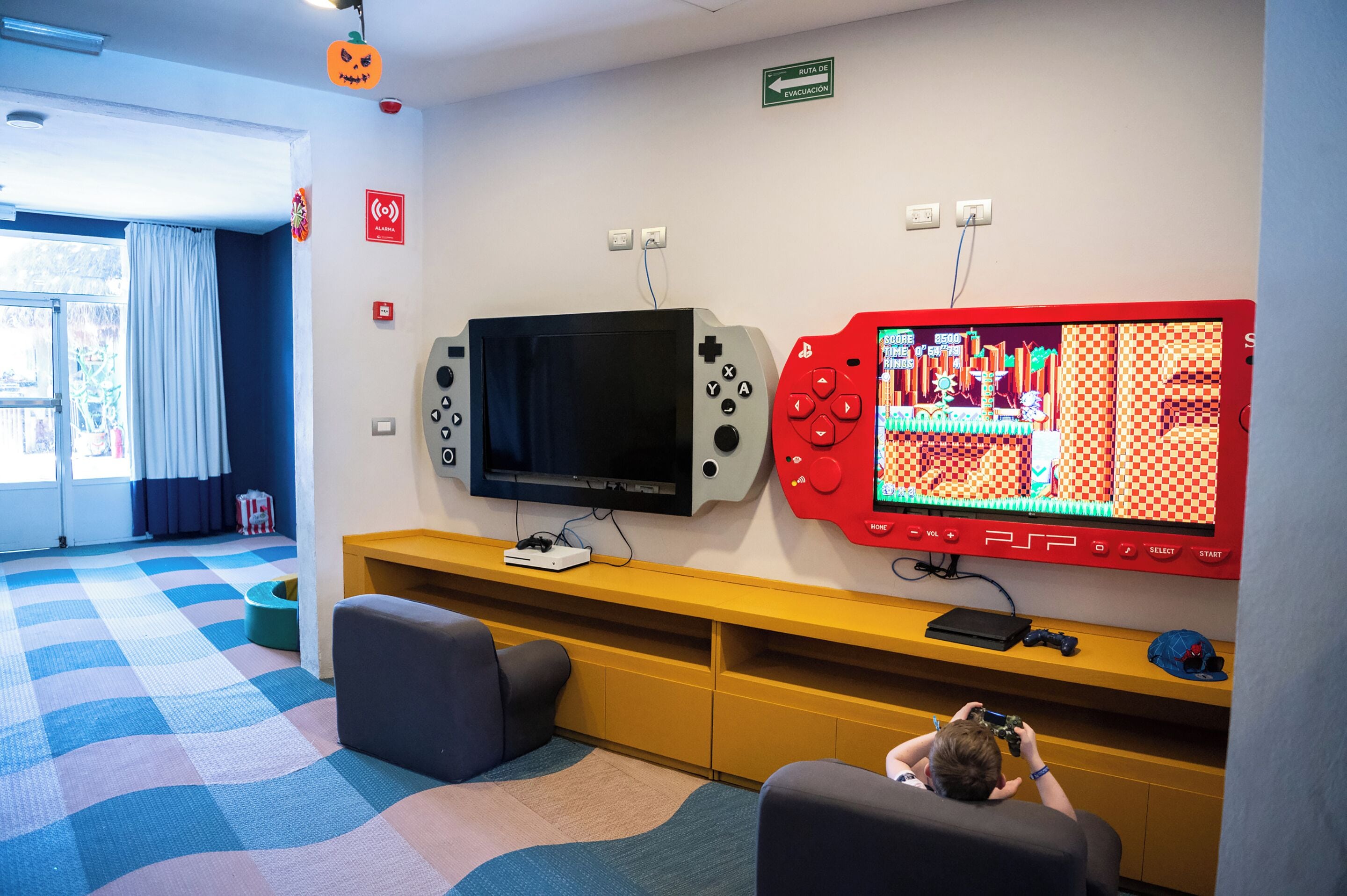Una habitación con suelo a cuadros, televisor, consola de videojuegos y una persona jugando.