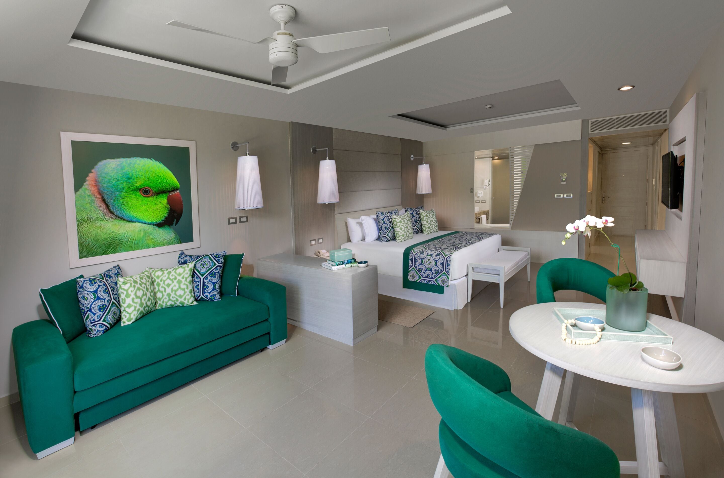 Une chambre d’hôtel moderne avec un canapé vert, un lit avec une literie à motifs, une petite table sur laquelle est posé un vase de fleurs et un ventilateur de plafond.