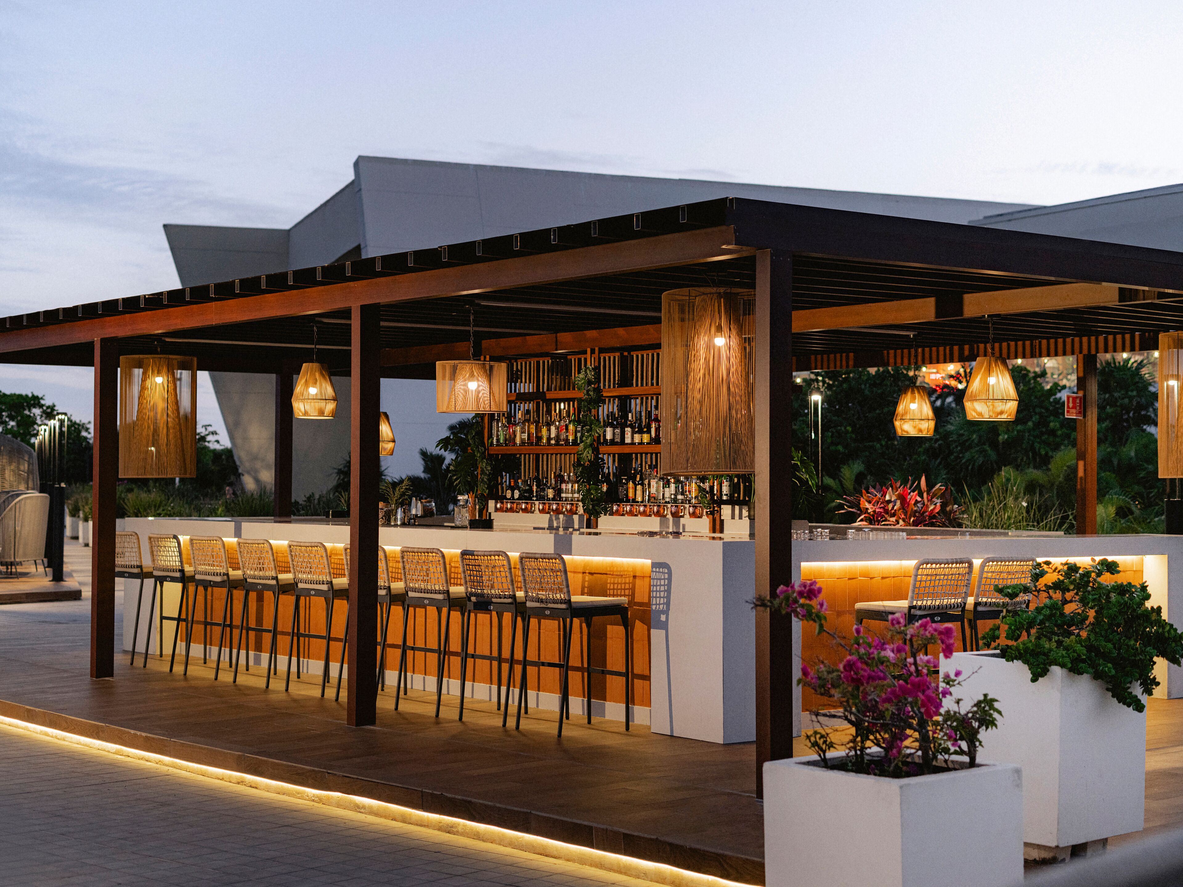 An outdoor bar area with a long counter, multiple chairs, and a variety of bottles and glasses on shelves.