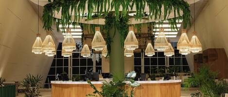 A reception area with a wooden reception desk, hanging wicker pendant lights, and a large central column with hanging greenery.