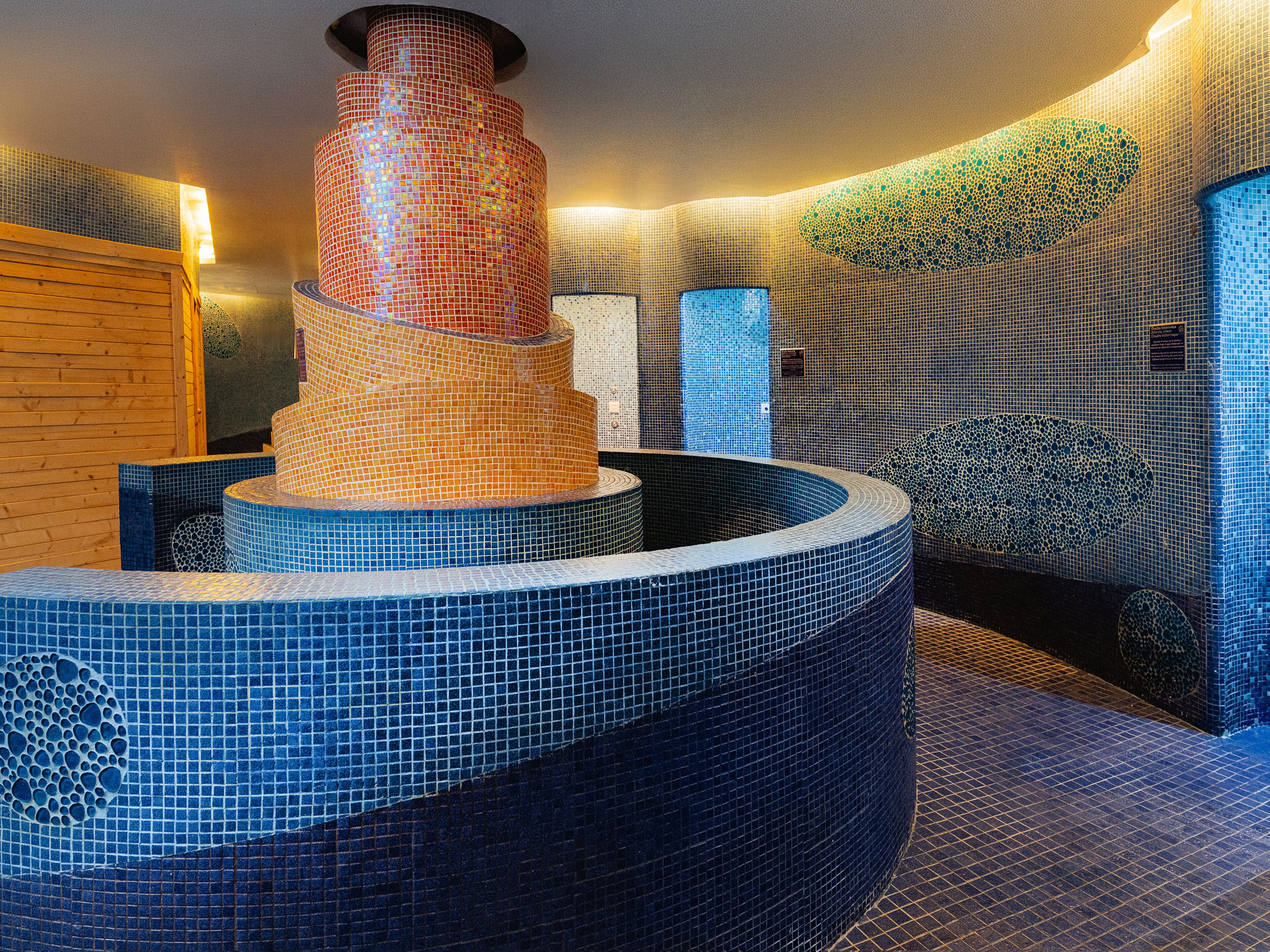 A modern spa area with a central mosaic fountain, tiled walls, and a wooden bench.