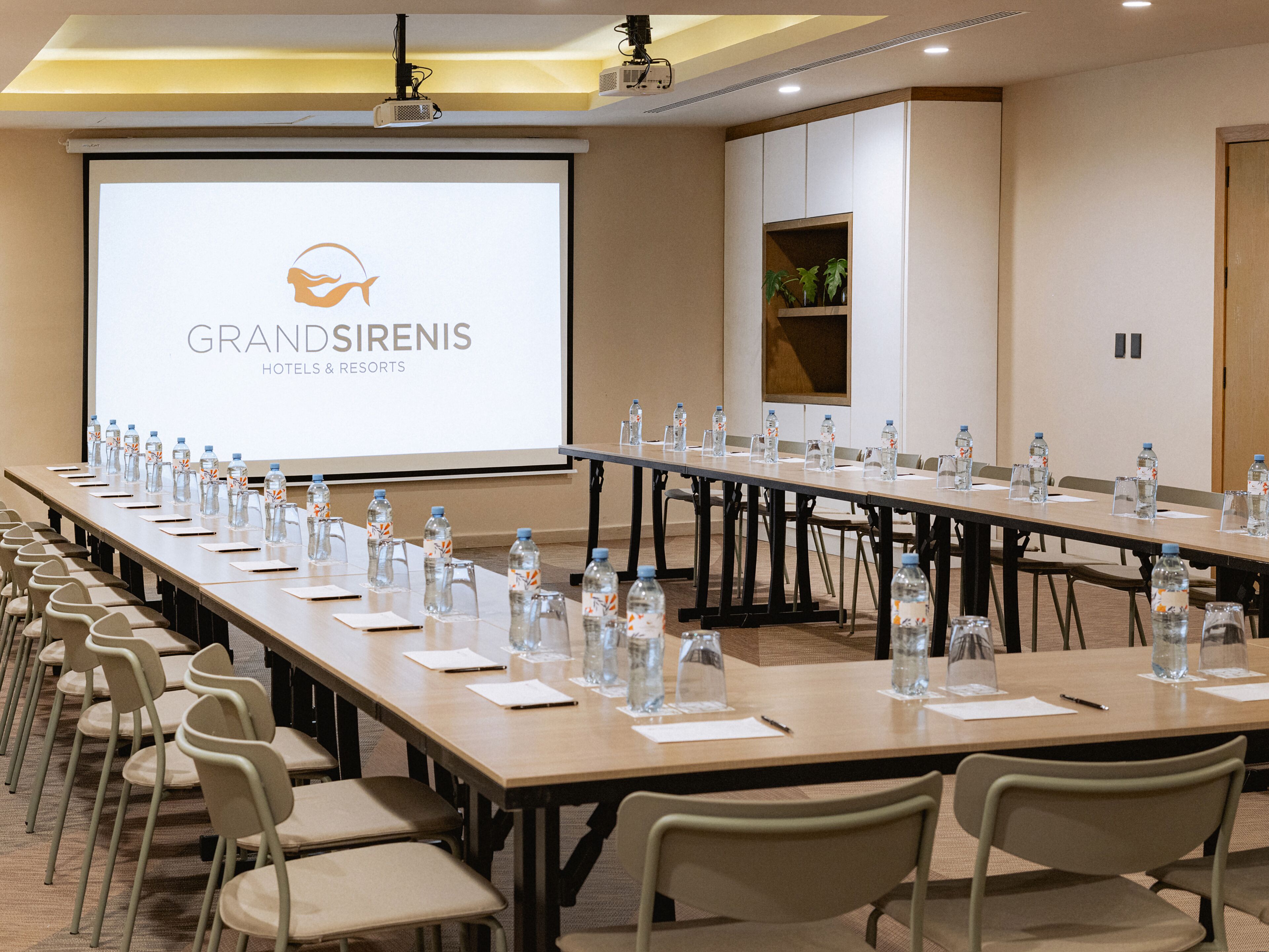 A conference room with a long table, chairs, water bottles, and a projector screen displaying the GrandSirenis Hotels & Resorts logo.