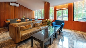 Un salon moderne avec un canapé en cuir, une table basse en verre et un fauteuil bleu. Les murs sont recouverts de lambris en bois et il y a une grande fenêtre avec des stores.