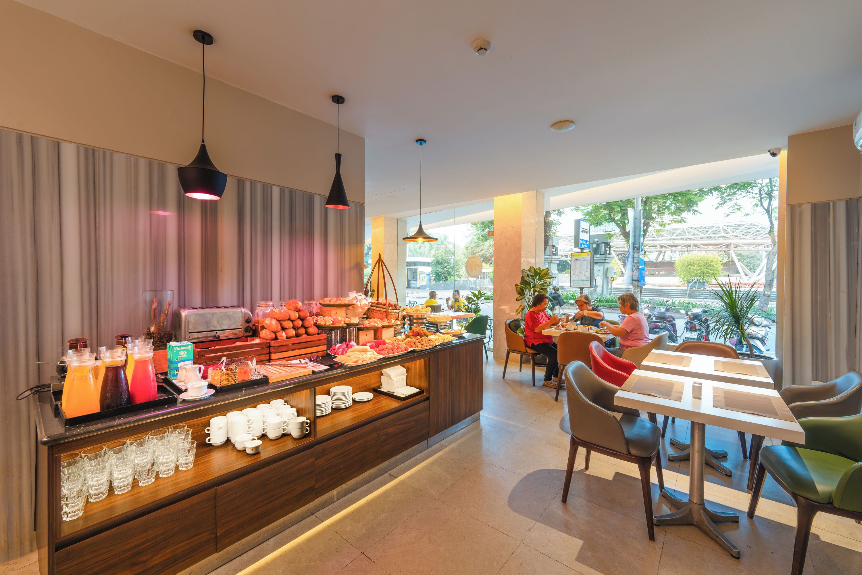 A breakfast buffet with various food items and beverages, a seating area with tables and chairs, and large windows offering a view of the outside.