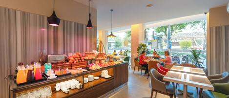 A breakfast buffet with various food items and beverages, a seating area with tables and chairs, and large windows offering a view of the outside.