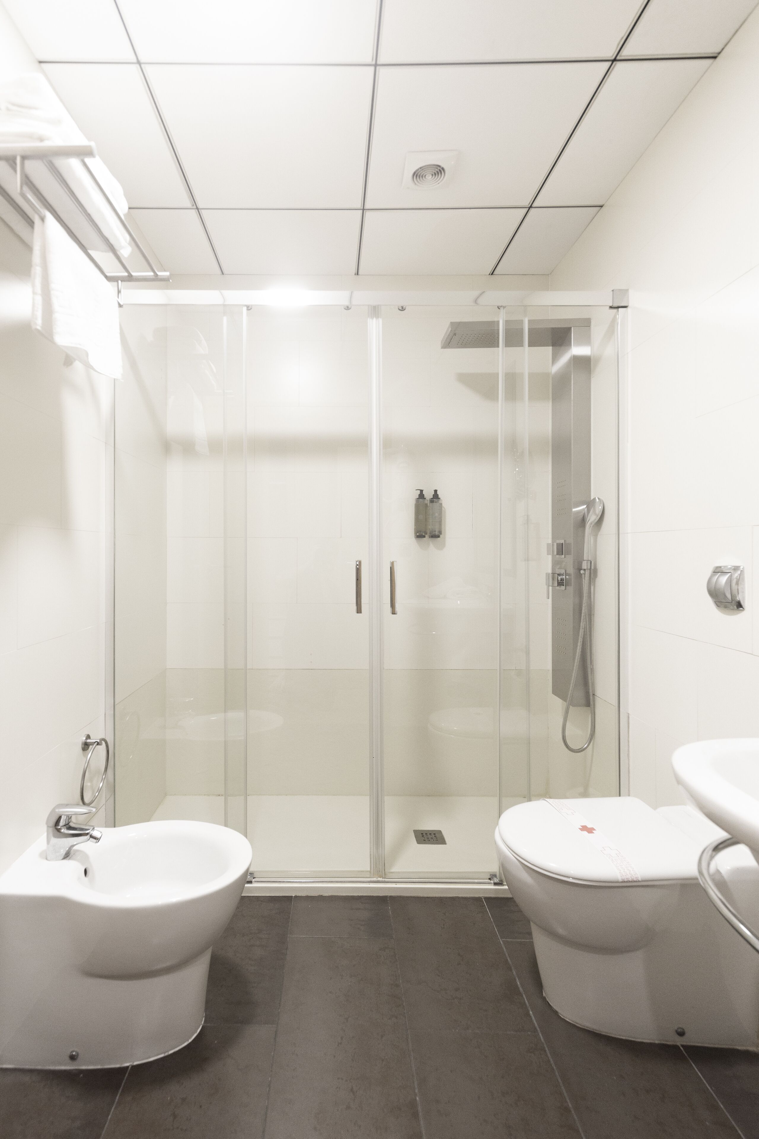 Un baño moderno con ducha de vidrio, dos inodoros blancos y un bidé.