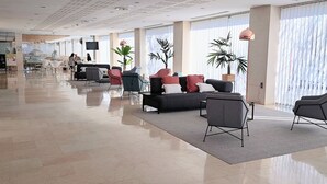Un hall d’entrée spacieux au design moderne, comprenant un espace détente avec des canapés et des fauteuils, de grandes fenêtres équipées de stores et un sol ciré.