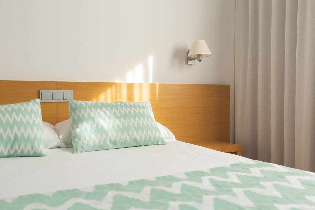 Una habitación de hotel con cama, dos almohadas, una mesita de noche, lámpara y ventana con cortinas.