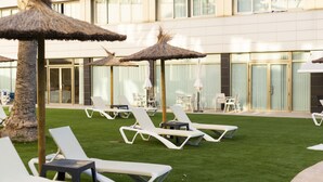 Une cour d’hôtel avec des chaises longues blanches, des parasols en chaume et un palmier.