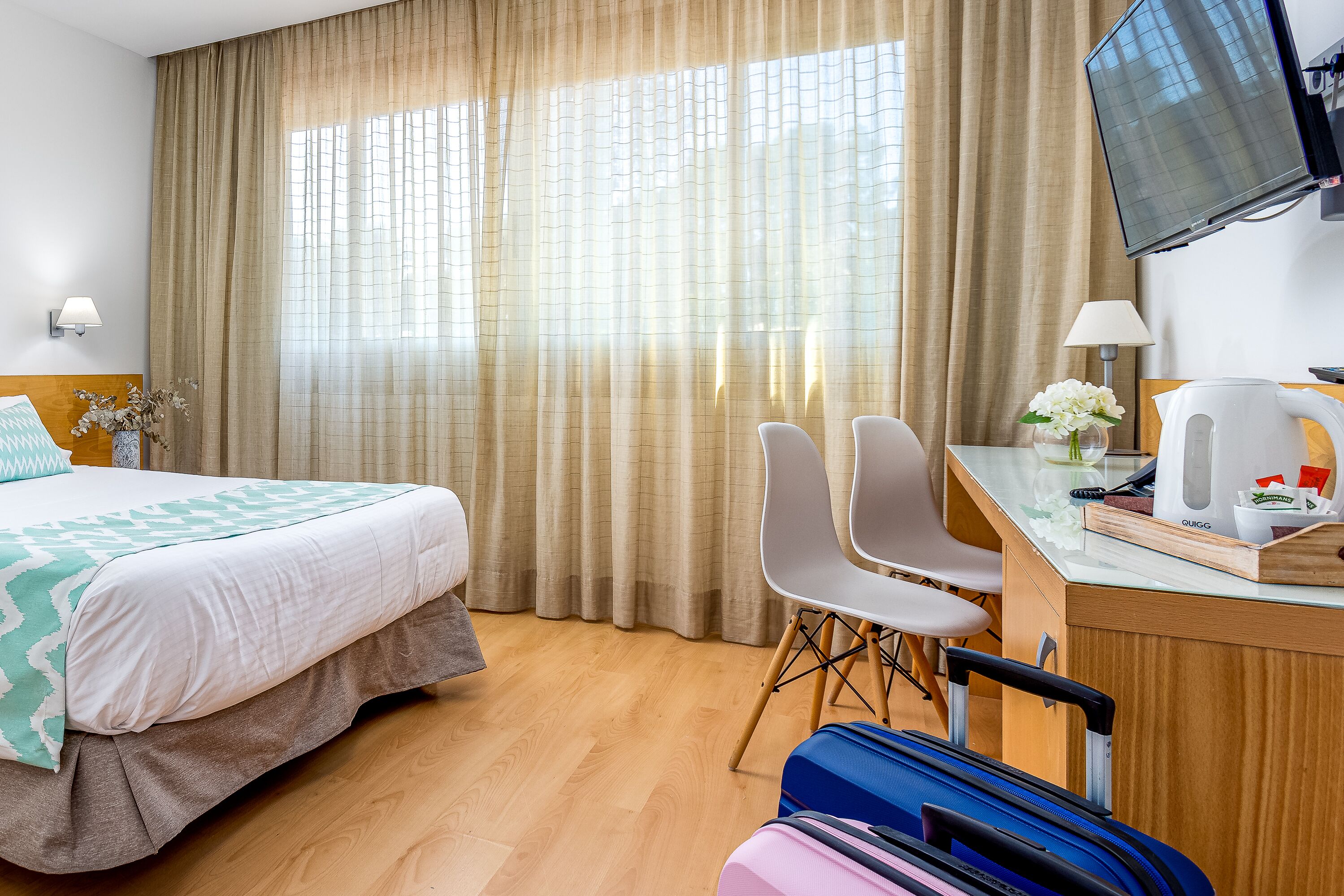 Una habitación de hotel con cama, escritorio con silla, televisor y ventana con cortinas.