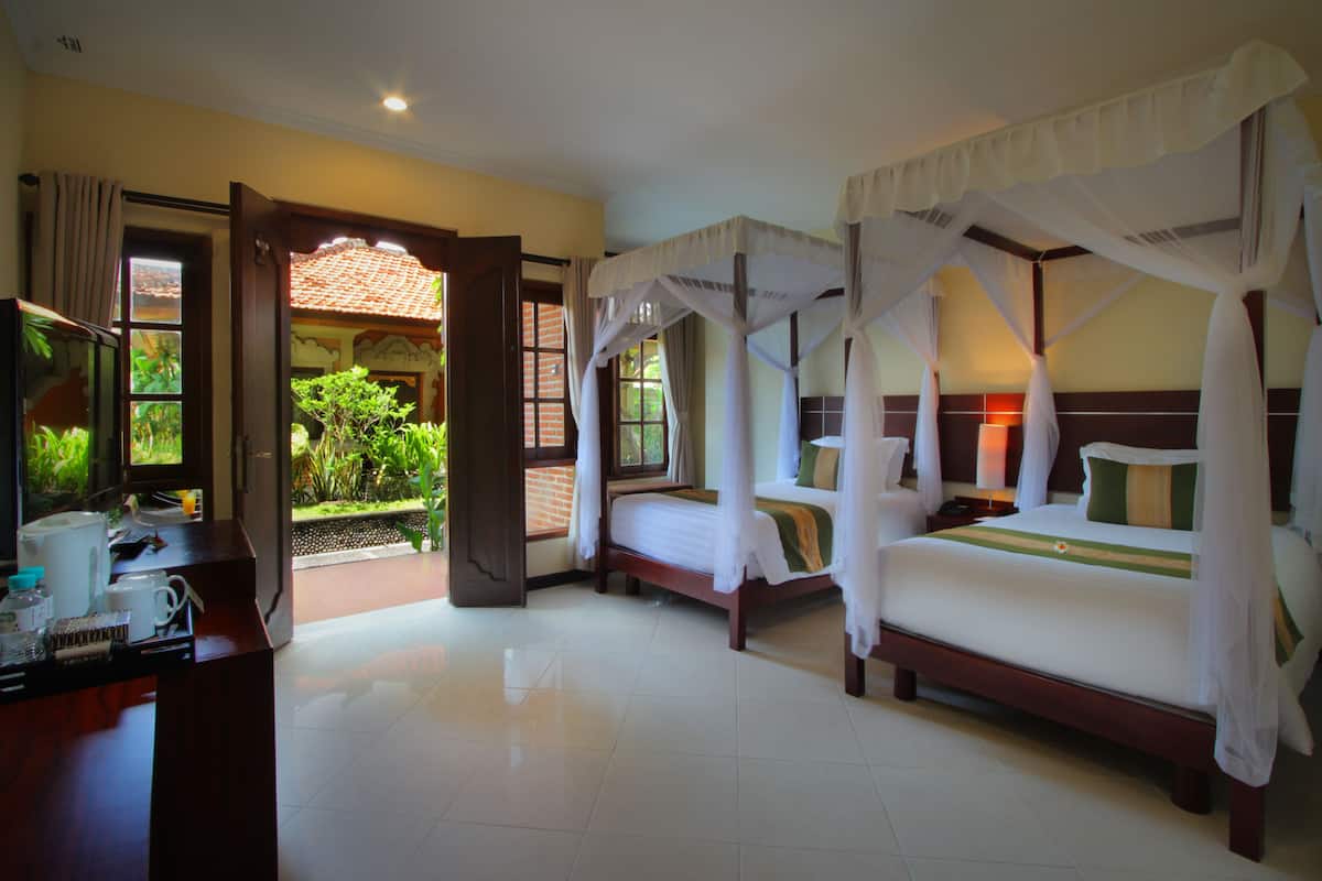 A hotel room with two beds, a desk, and a view of a garden through an open door.