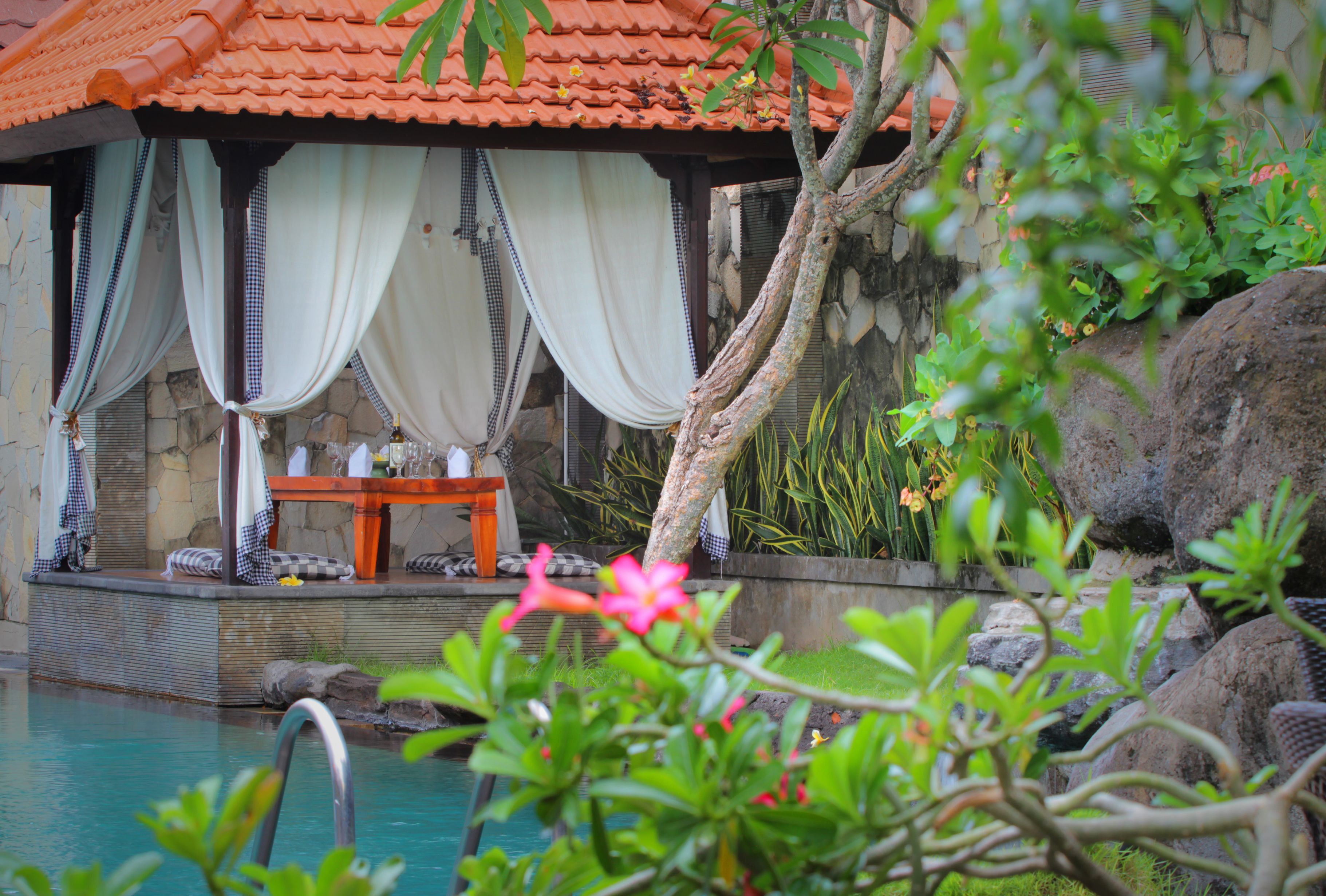 A pool area with a stone wall, a wooden deck, and a covered seating area with a table and chairs.