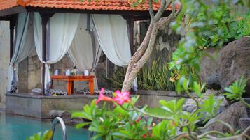 A pool area with a stone wall, a wooden deck, and a covered seating area with a table and chairs.