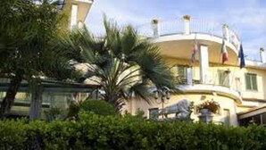 A large, multi-story building with a curved facade, surrounded by lush greenery and palm trees. - Hotel Leonessa (Volla)