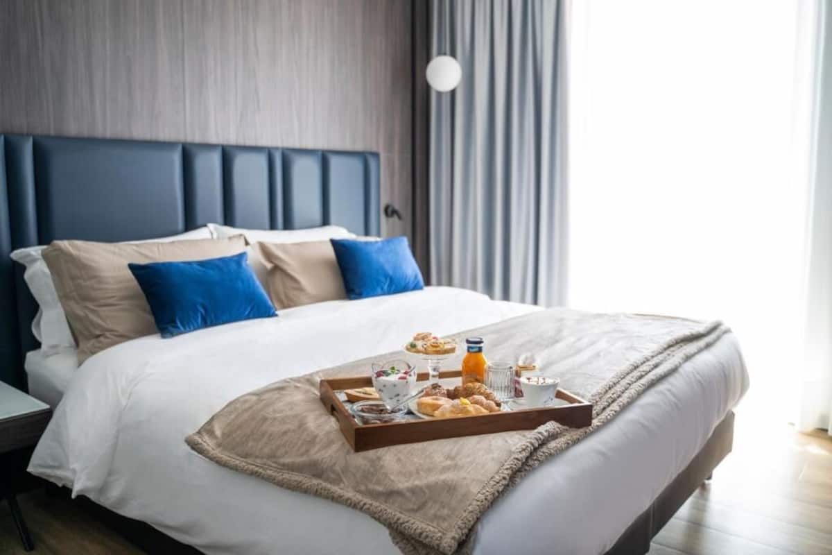 A neatly made bed with a tray of breakfast items, including pastries and a drink, in a modern hotel room with a grey headboard and blue pillows.