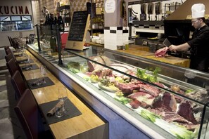 A butcher section with various meats displayed in a glass case, a chef preparing meat, and a chalkboard menu in the background. - Hotel Leonessa (Volla)
