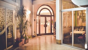 Un couloir d’hôtel avec une entrée constituée d’une porte vitrée, des plantes en pot et un espace détente avec des tables et des chaises.