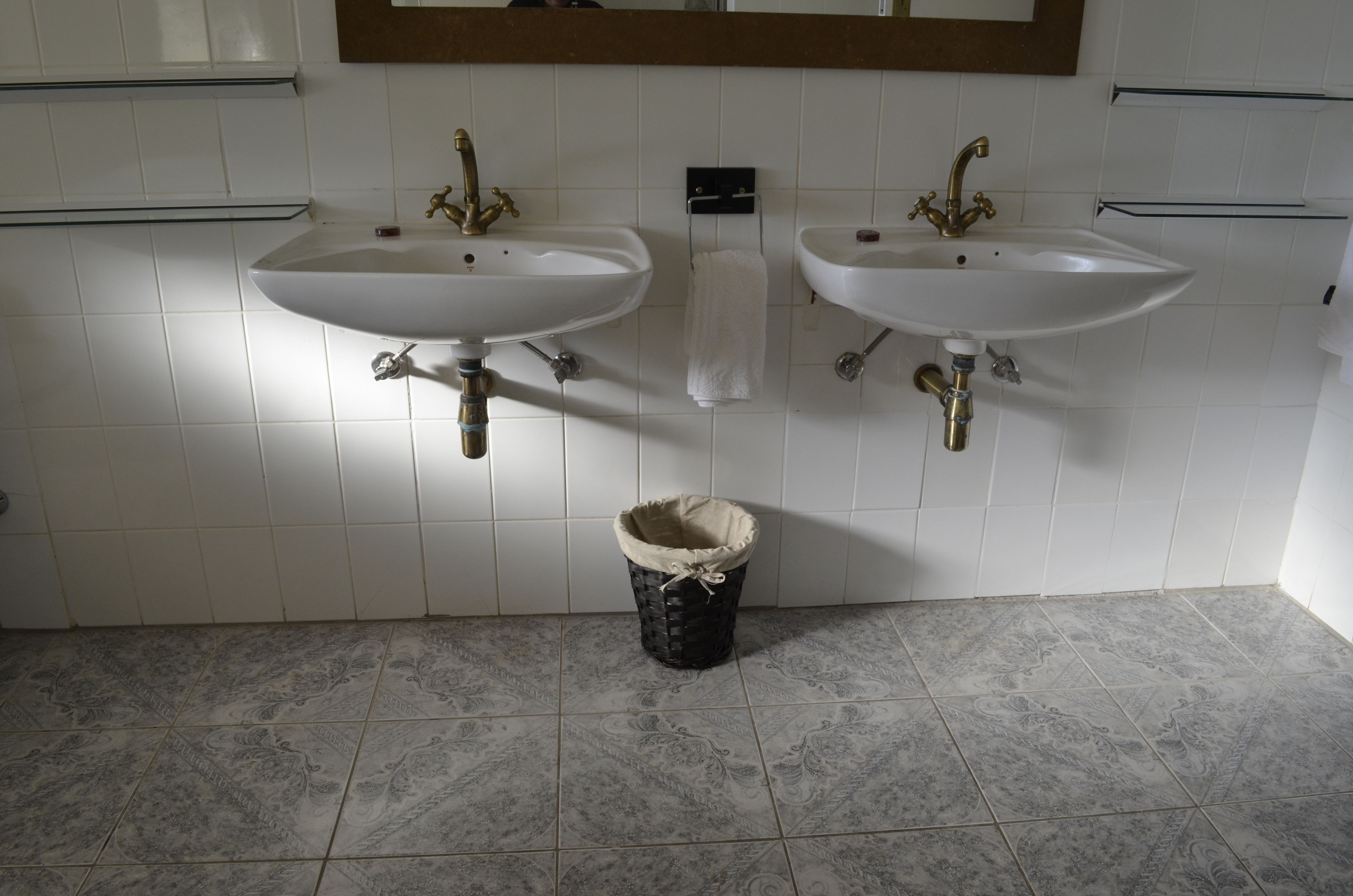 Un baño con dos lavamanos blancos, un espejo encima y un cubo de basura adelante.