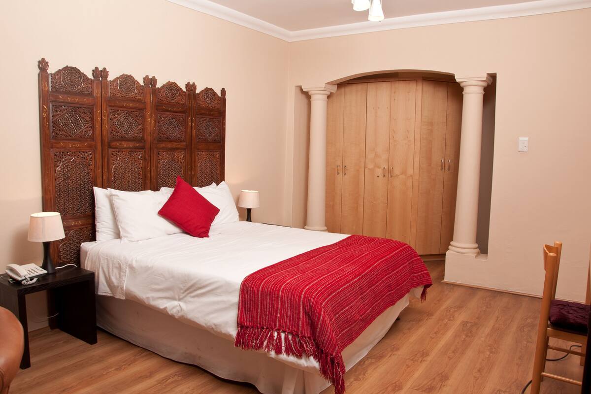 A neatly made bed with white linens and a red throw, a wooden headboard, a nightstand with a lamp, and a wooden wardrobe.