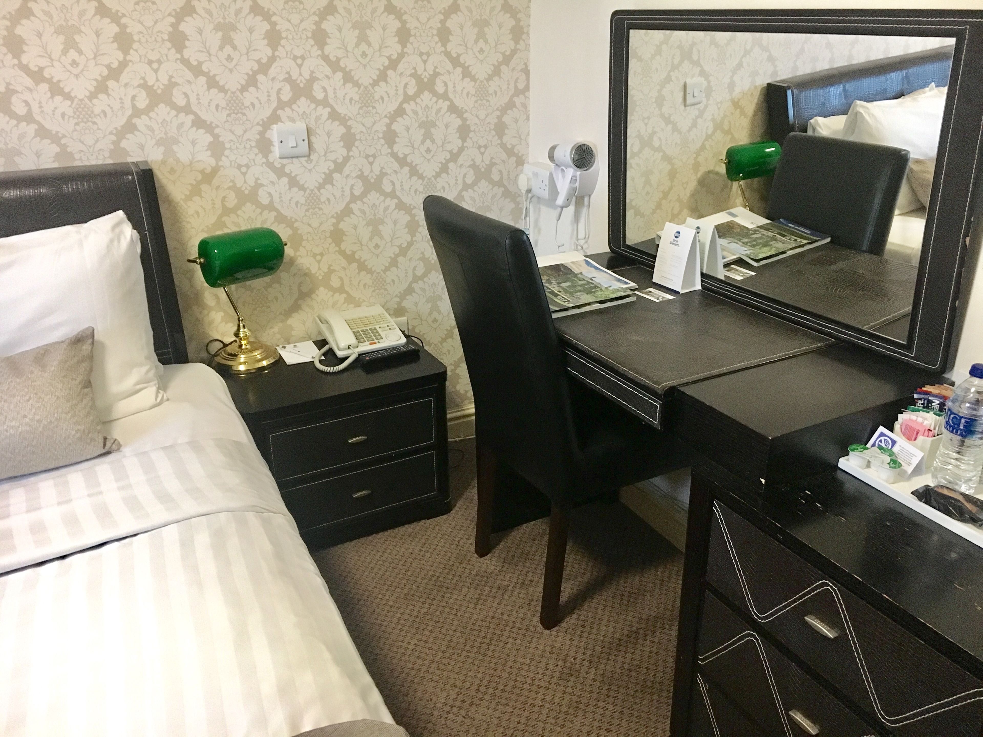 A hotel room with a bed, a desk with a telephone, a chair, a mirror, and a nightstand with a lamp.