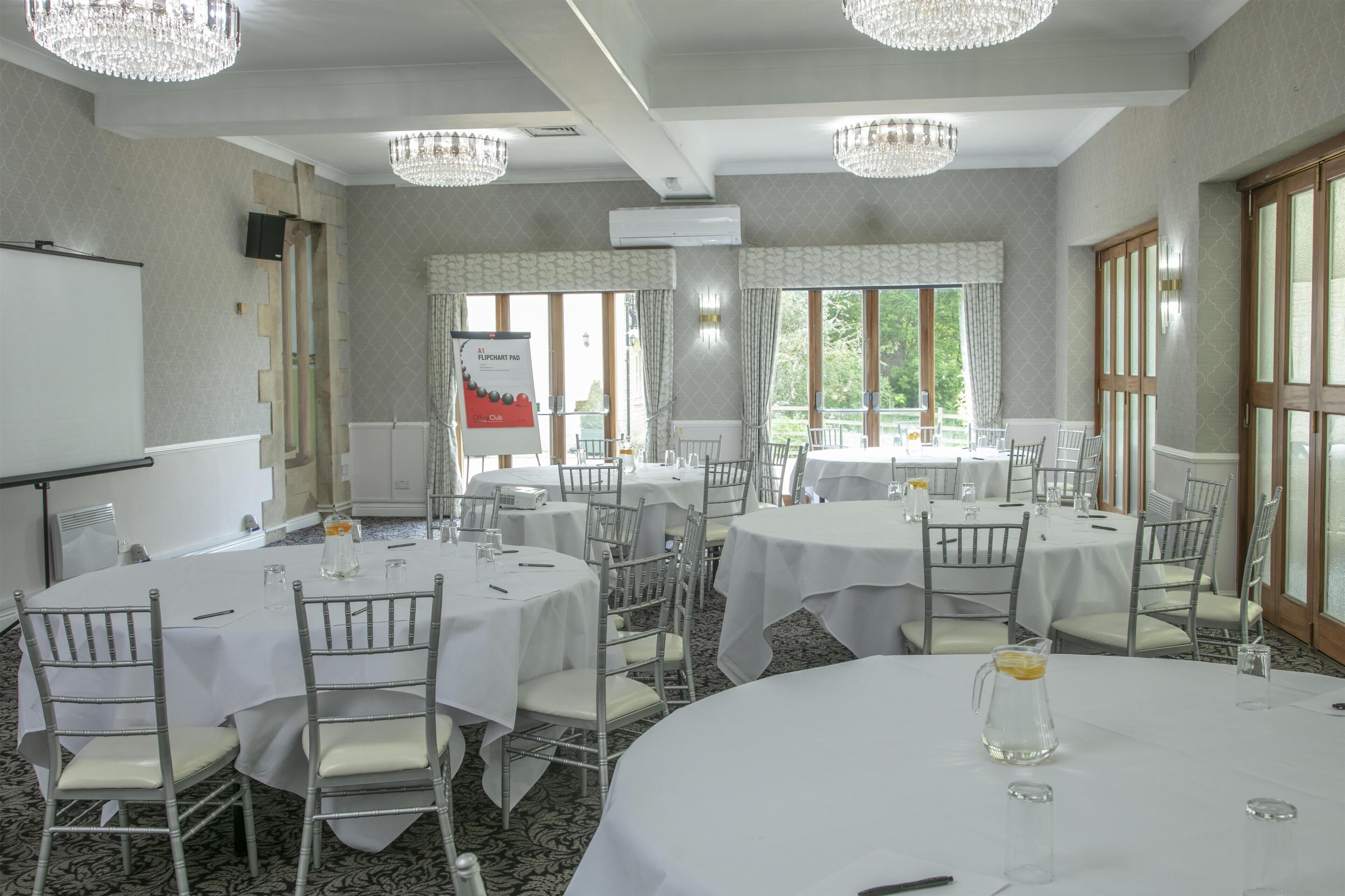 Un salón de conferencias con varias mesas redondas, manteles blancos y sillas plateadas. Hay lámparas de araña colgando del techo y grandes ventanales con cortinas.