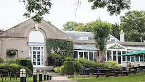 A building with a large arched entrance, a covered patio area with outdoor seating, and a gravel pathway leading to the entrance. - Normanton Park Hotel, BW Signature Collection (Oakham)