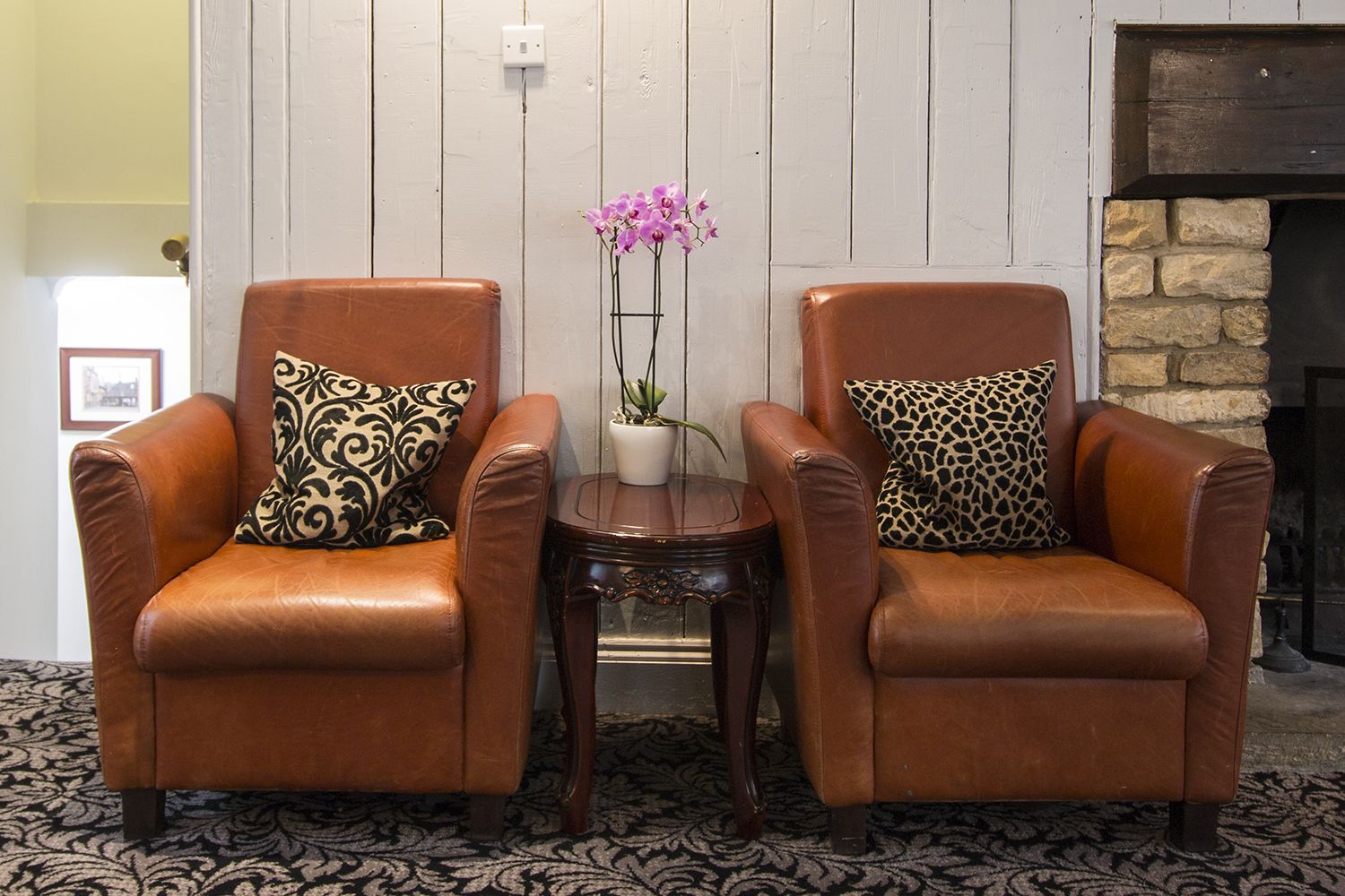 Twee bruine leren fauteuils met gestreepte kussens, een bijzettafeltje met een potorkidee en een stenen openhaard op de achtergrond.